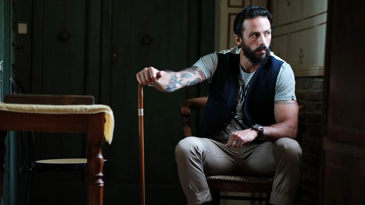 Homme barbu en gilet, assis près d’une table de billard dans un intérieur aux tons profonds.